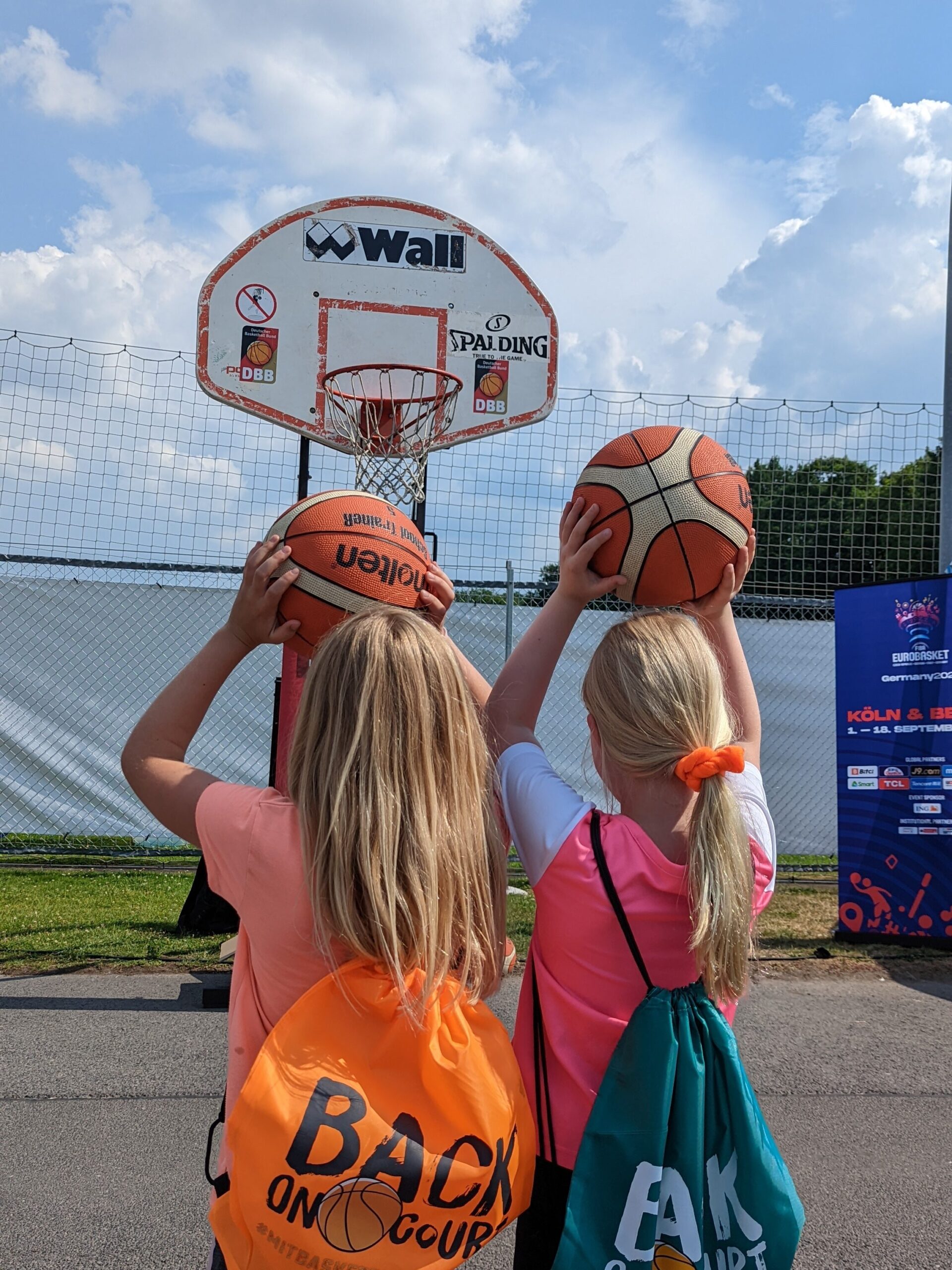 zwei Mädchen mit bunten Turnbeuteln stehen vor dem Basketballkorb
