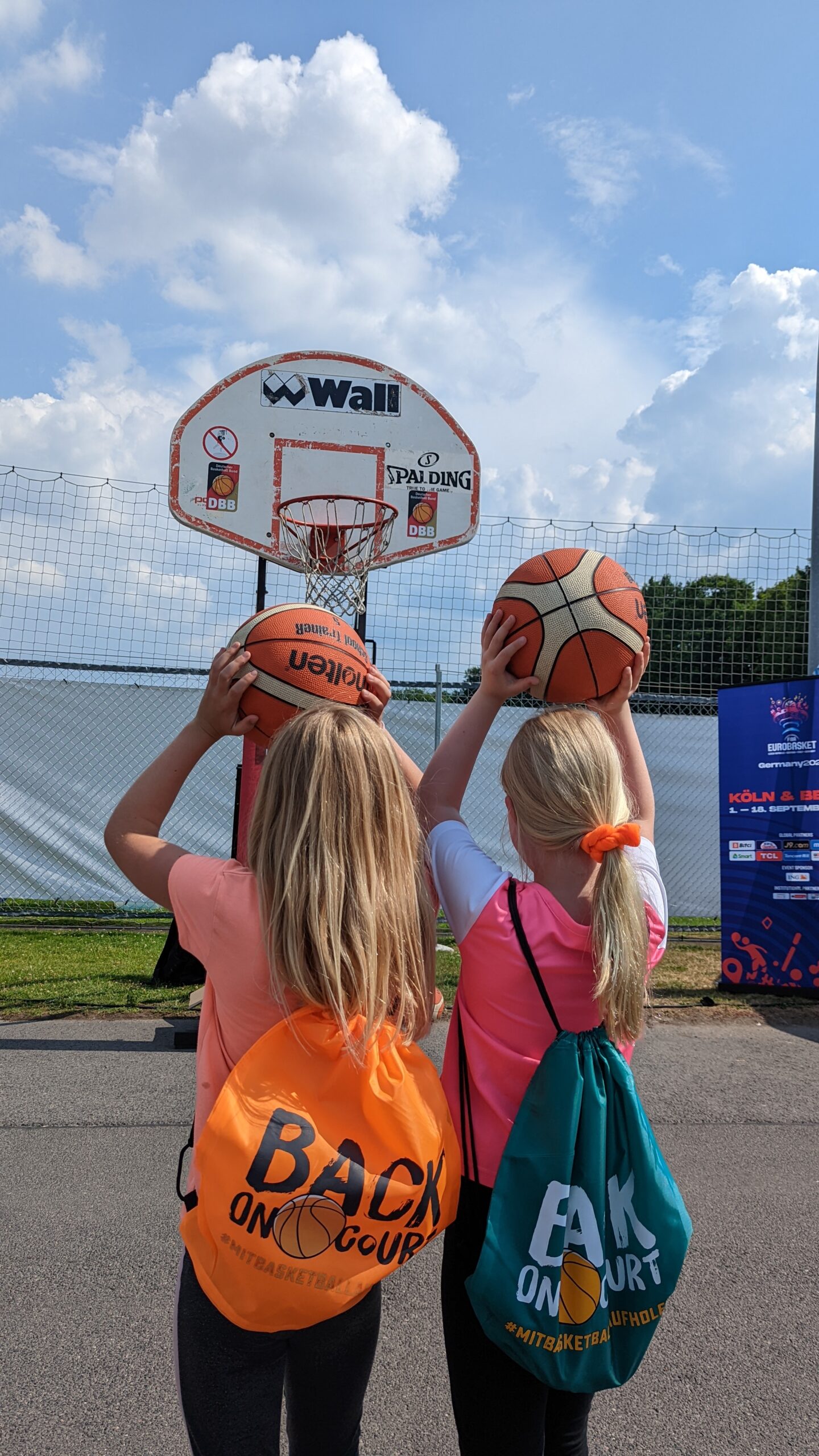 zwei Mädchen mit bunten Turnbeuteln stehen vor dem Basketballkorb